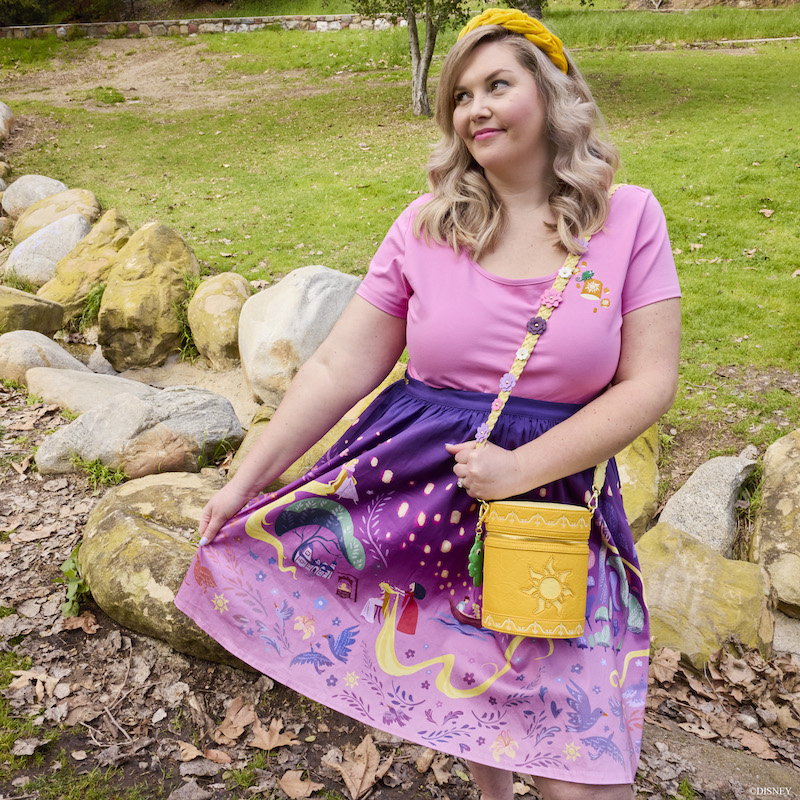 Woman outside wearing the Rapunzel Sandy Skirt, holding out one side to feature the artwork of Rapunzel with Mother Gothel. She's also wearing the pink Rapunzel Lanterns Kelly Top and the figural lantern crossbody